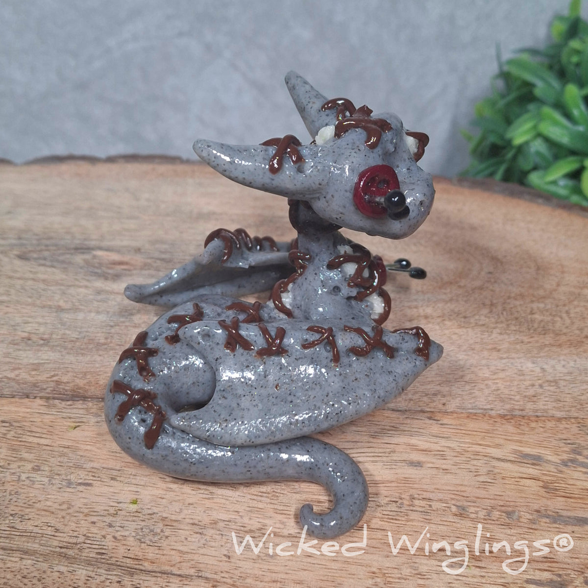 A clay voodoo dragon on a wood surface with greenery. Text says Wicked Winglings