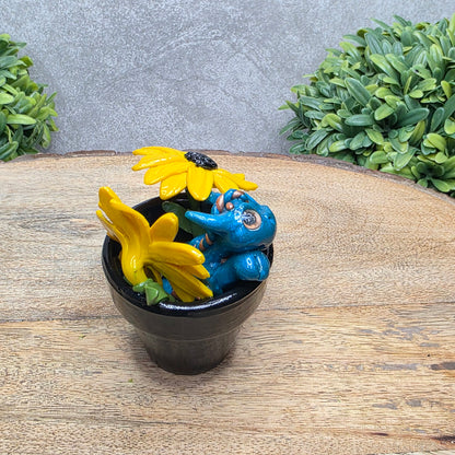 Side view of a clay baby dragon in a pot with black eye susans against a wood surface with greenery.