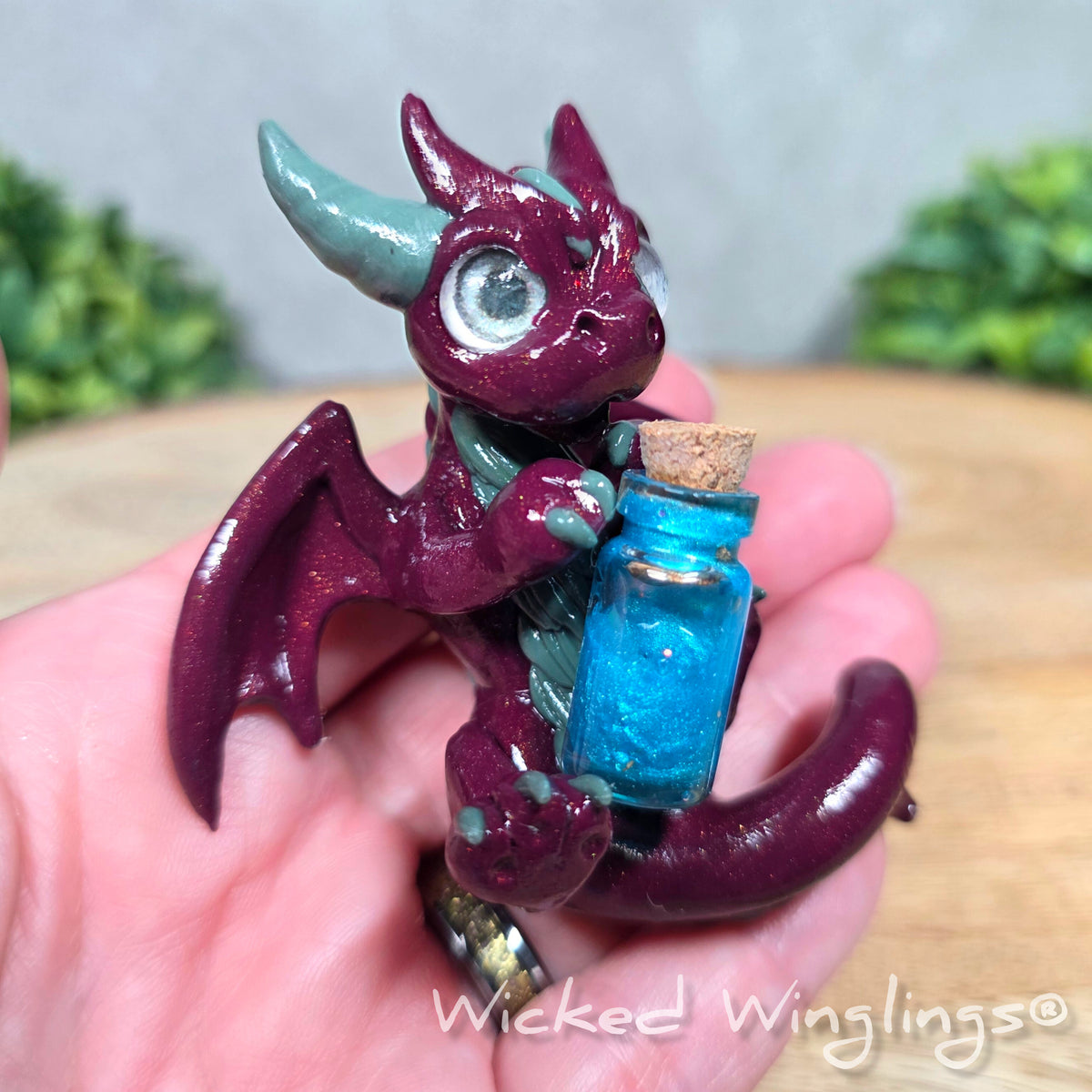 A hand holding a clay dragon with a potion bottle. Text reads Wicked Winglings.
