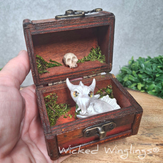 A hand holding a clay dragon sitting in a chest accented with moss and a skull. Text says Wicked Winglings