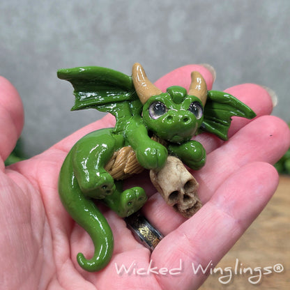 A hand holding a clay dragon with a skull on a wood surface with greenery. Includes text Wicked Winglings
