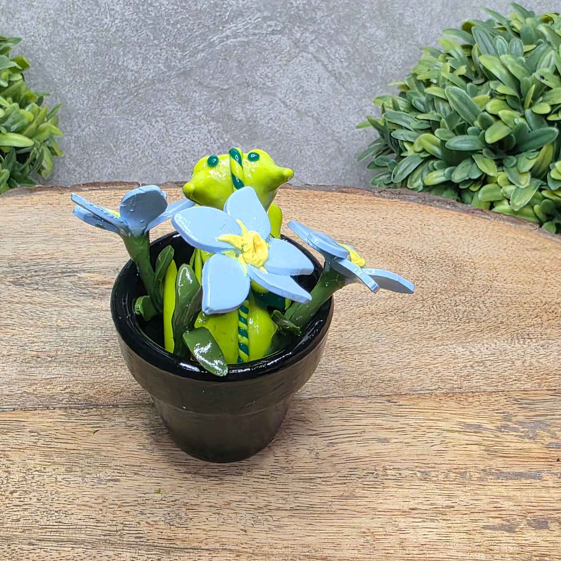 Back view of a clay baby dragon in a pot with forget me nots against a wood surface with greenery.