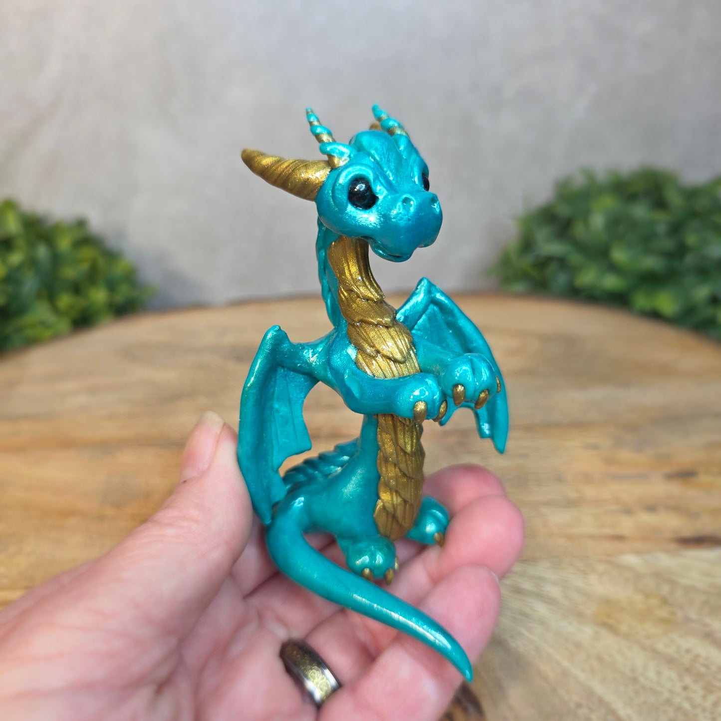 A hand holding a teal and gold clay dragon against a blurred wood surface