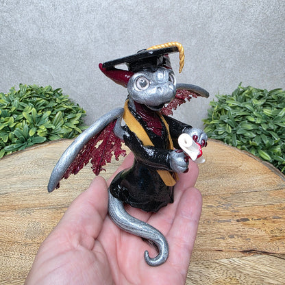 A hand holding a clay dragon in a graduation cap and gown holding a diploma