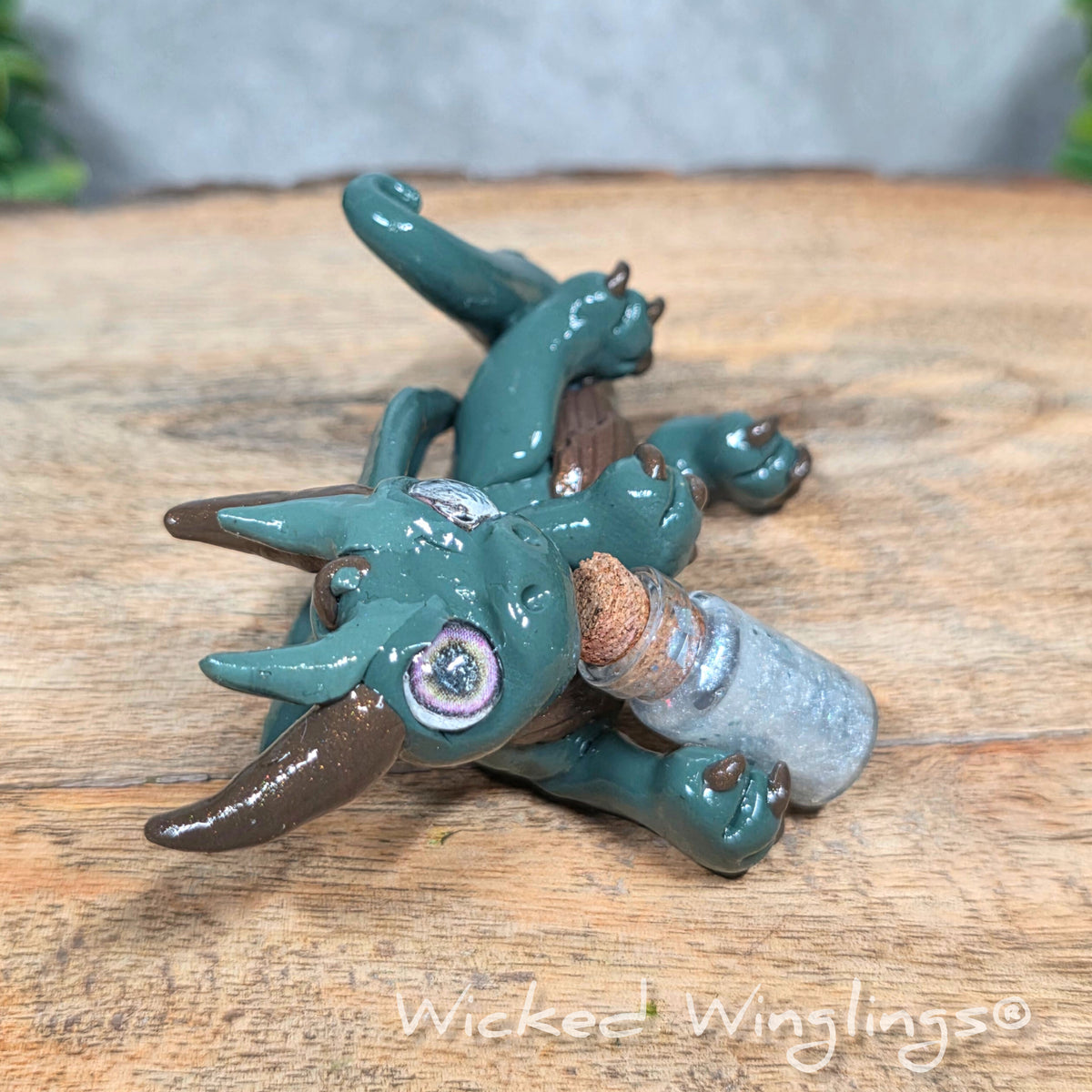 A clay dragon with a potion bottle on a wood surface. Text reads Wicked Winglings.