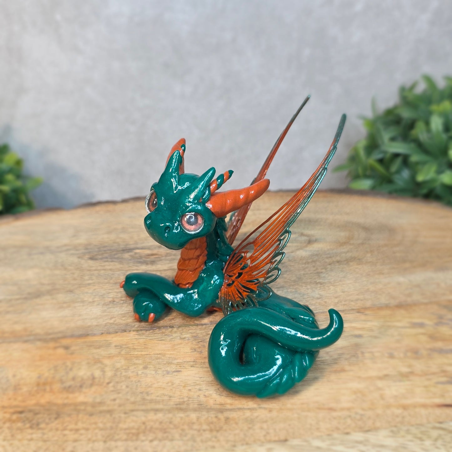 Green and orange dragon figurine on a wooden surface with a blurred green plant in the background