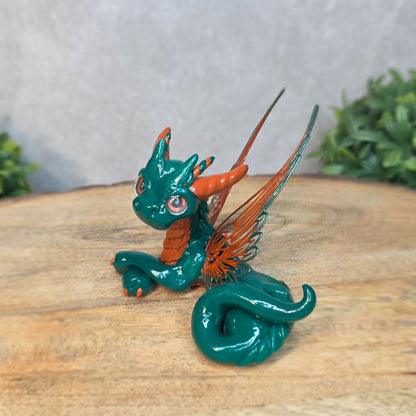 Green and orange dragon figurine on a wooden surface with a blurred green plant in the background