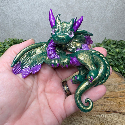 A hand holding a clay dragon against a wood background with greenery