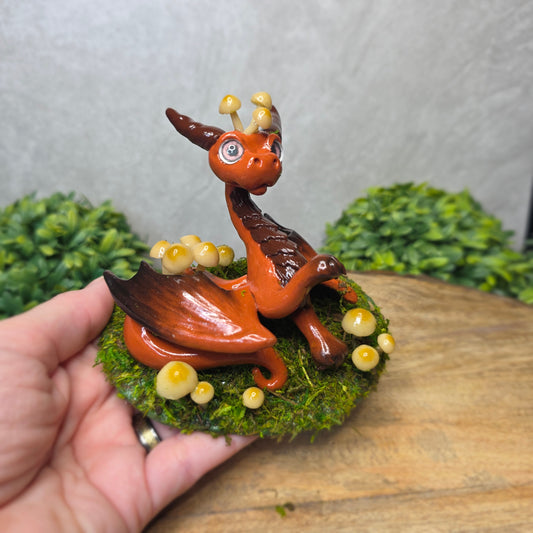 Hand holding a small dragon figurine on a bed of green moss with small yellow mushrooms