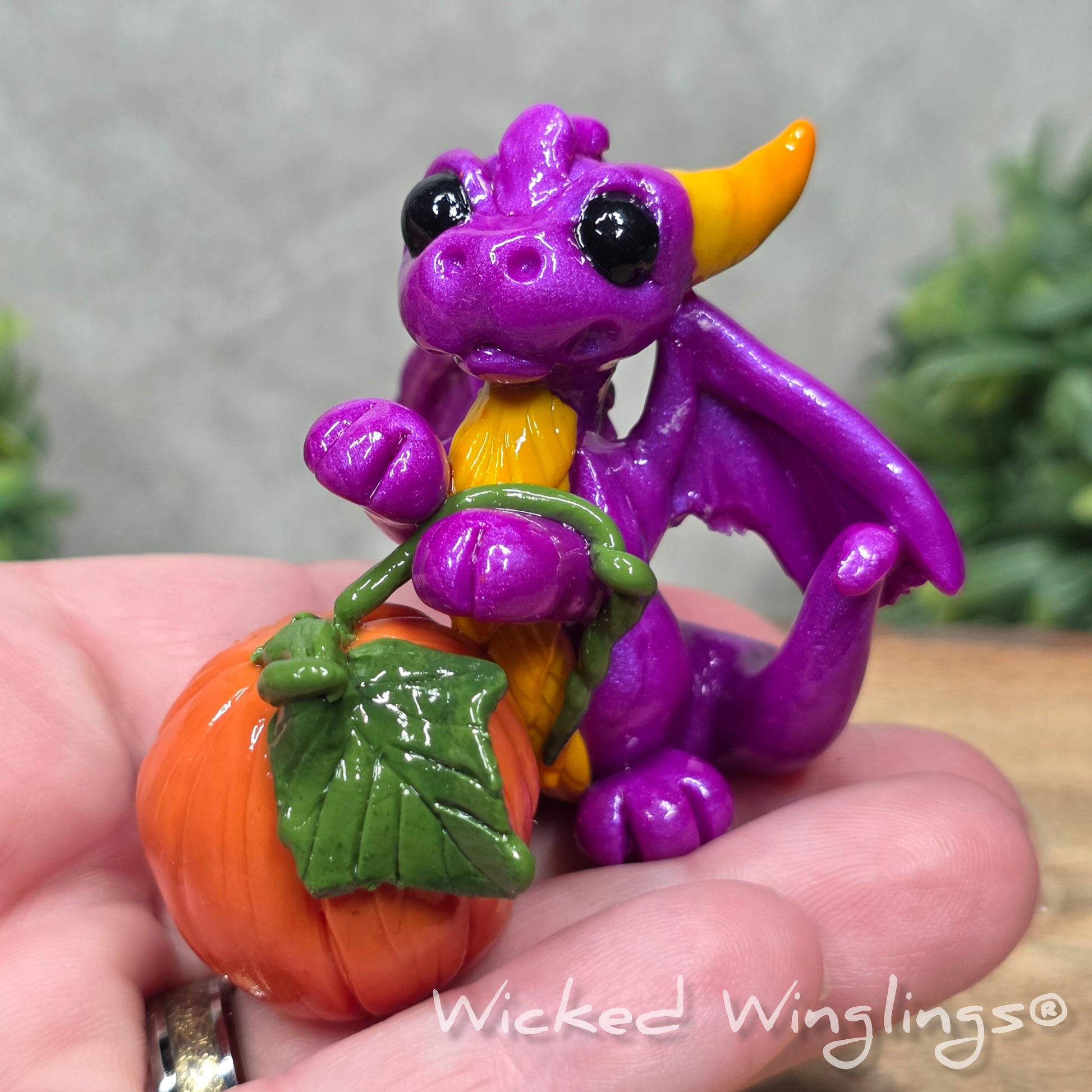 A hand holding a baby clay dragon sculpture with a pumpkin against a wood background with greenery and text Wicked Winglings