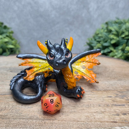 A clay dragon with a D20 dice against a wood surface with greenery.