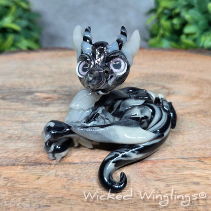 A swirled clay dragon in a wood stump against a blurred background with greenery. Text says Wicked Winglings