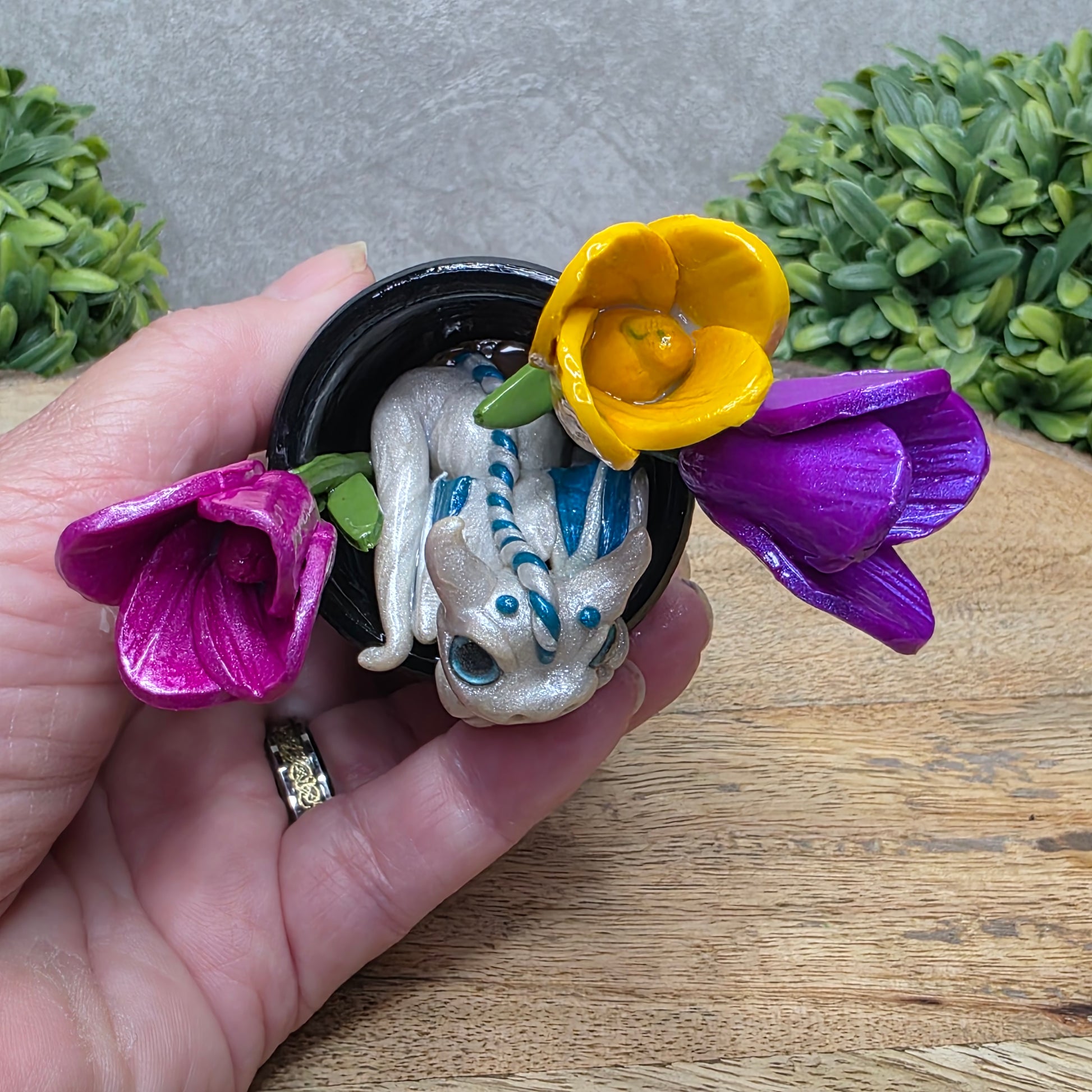 Top view of a clay baby dragon in a pot with tulips against a wood surface with greenery.
