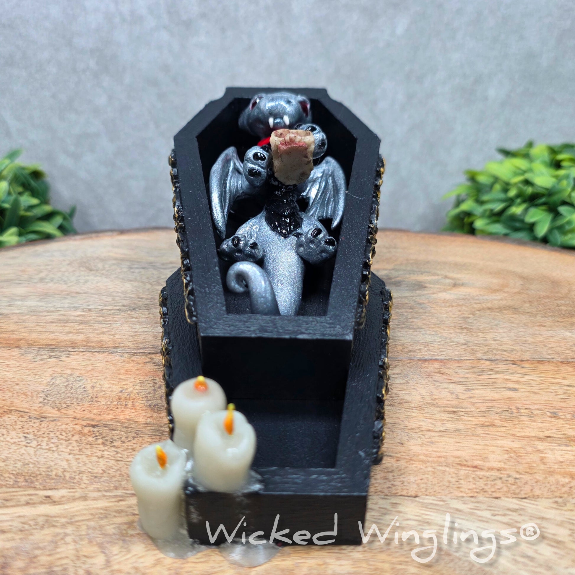 A clay dragon in a black wood coffin with candles and a skull on a wood surface with greenery. Includes text Wicked Winglings