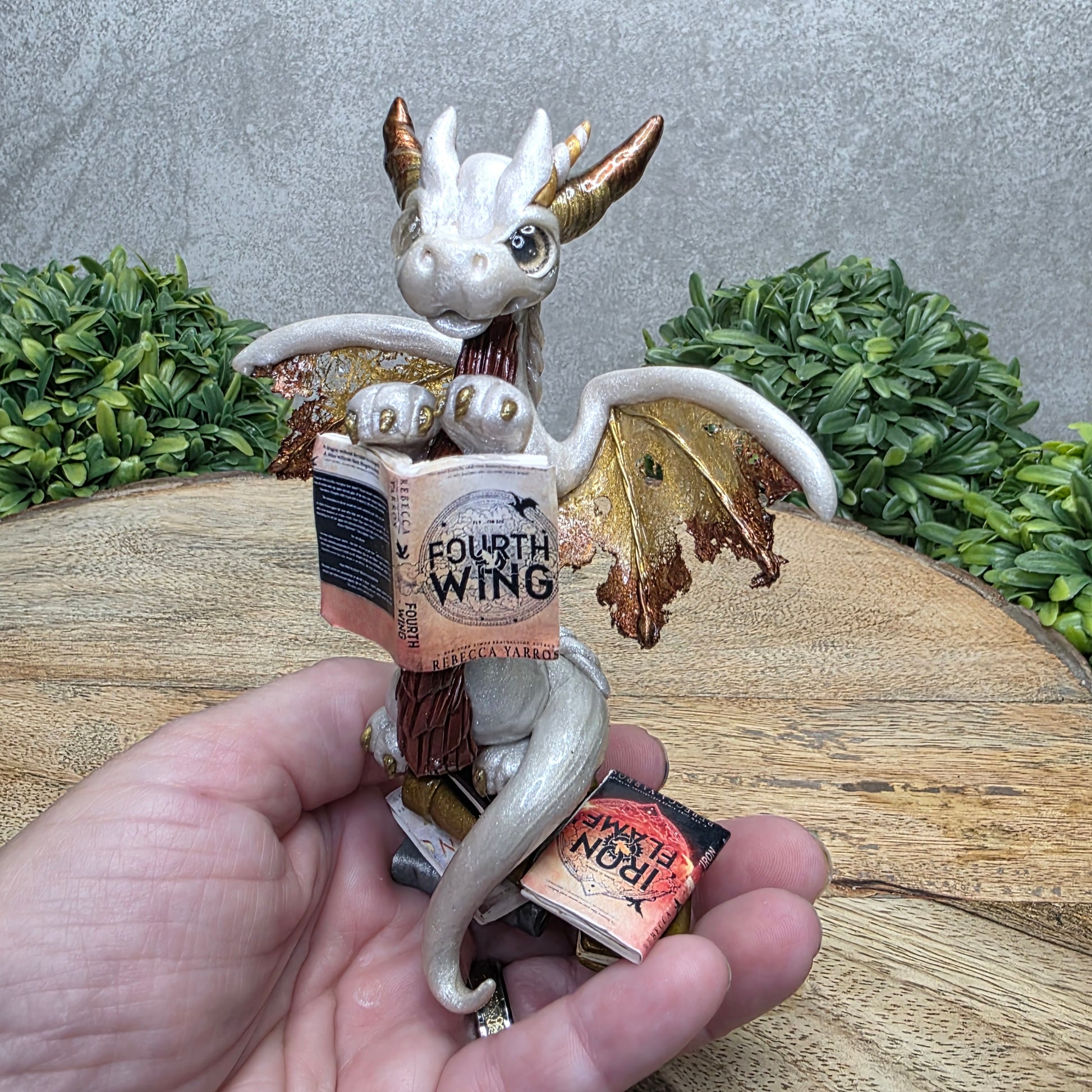 Hand holding a dragon reading a book against a wood surface with greenery