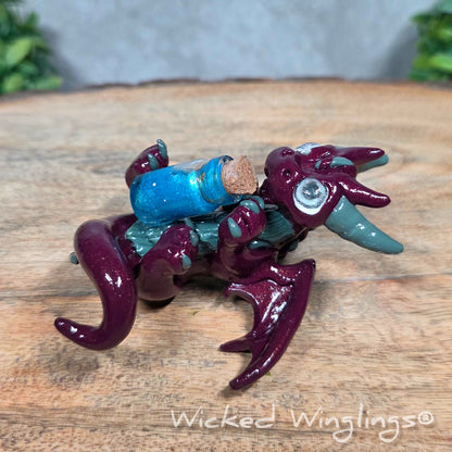 A clay dragon with a potion bottle on a wood surface. Text reads Wicked Winglings.