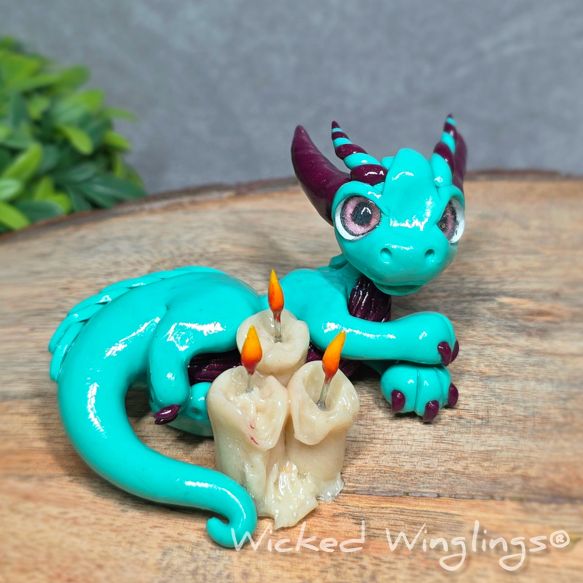 A clay dragon with candles against a wood surface with greenery. Text reads Wicked Winglings.
