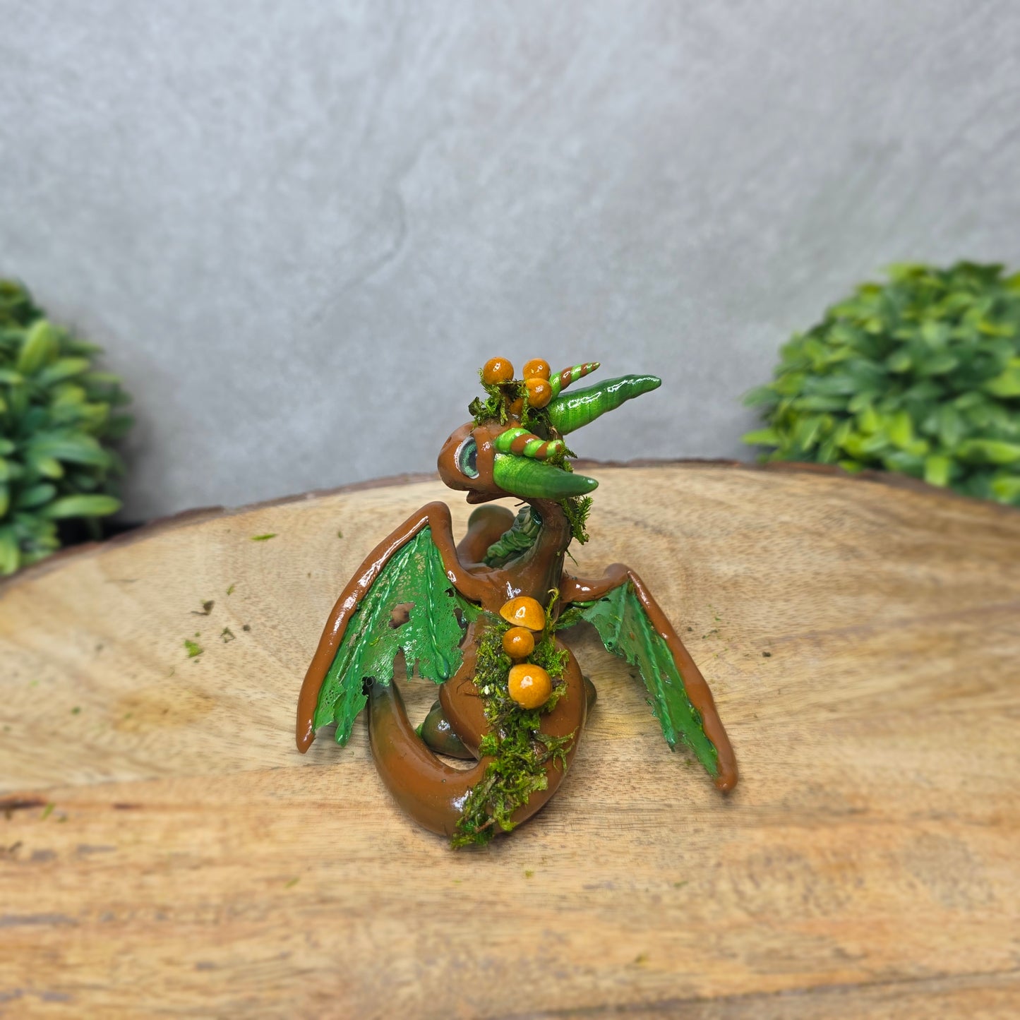 A clay dragon accented with moss and mushrooms against a wood surface with greenery.
