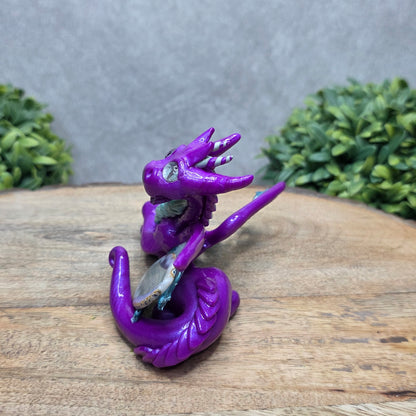 Side view of a clay dragon with an agate slice against a wood surface with greenery.