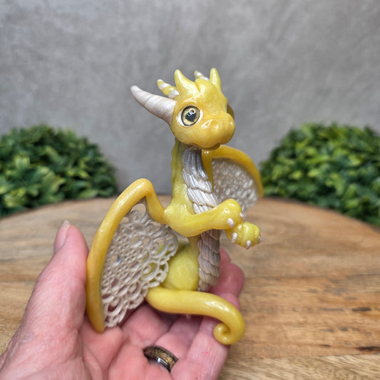 A hand holding a clay dragon sculpture with lacy wings on a wood surface with greenery.