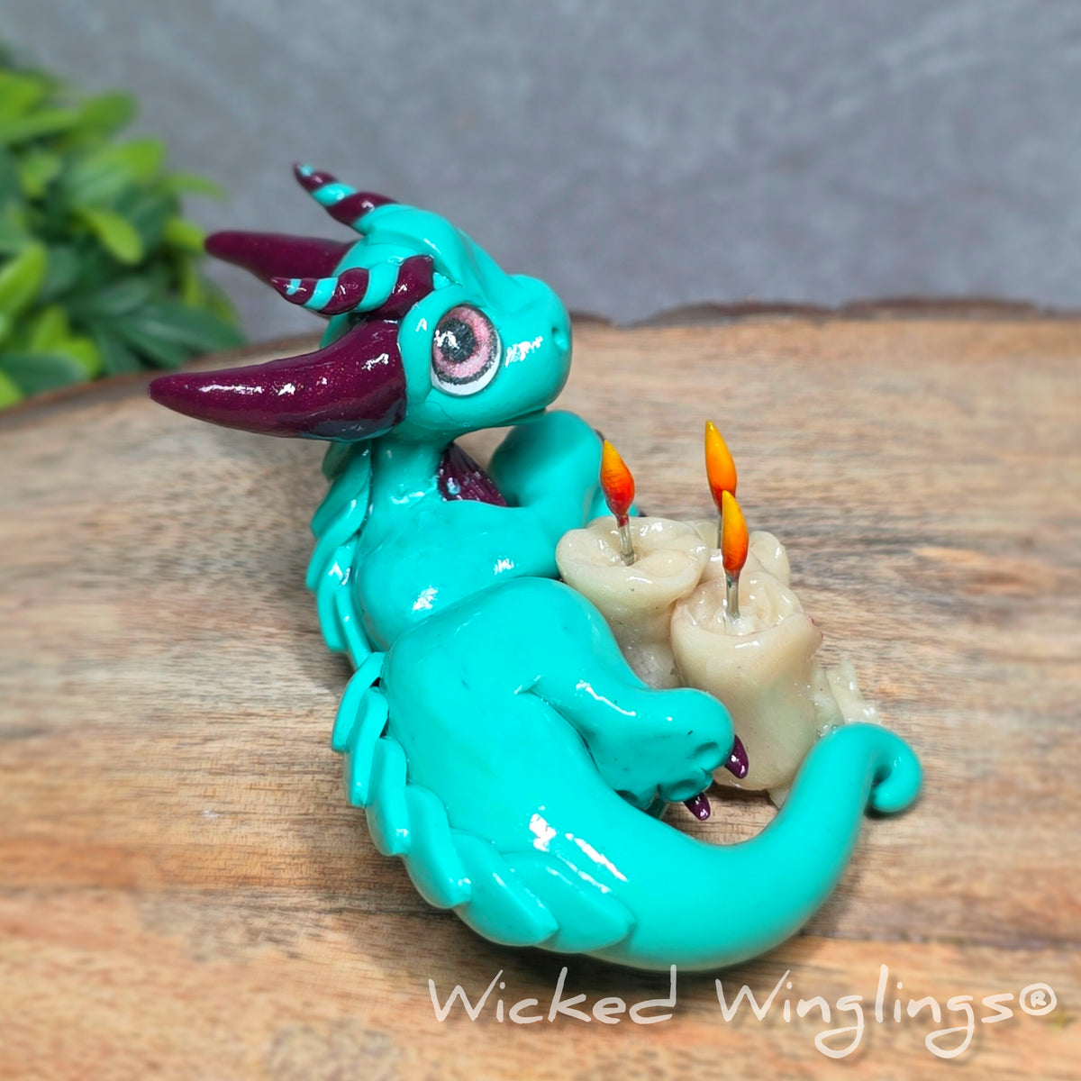 Side view of a clay dragon with candles against a wood surface with greenery. Text reads Wicked Winglings.