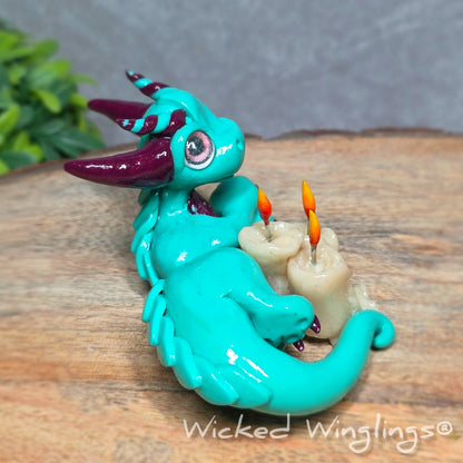 Side view of a clay dragon with candles against a wood surface with greenery. Text reads Wicked Winglings.