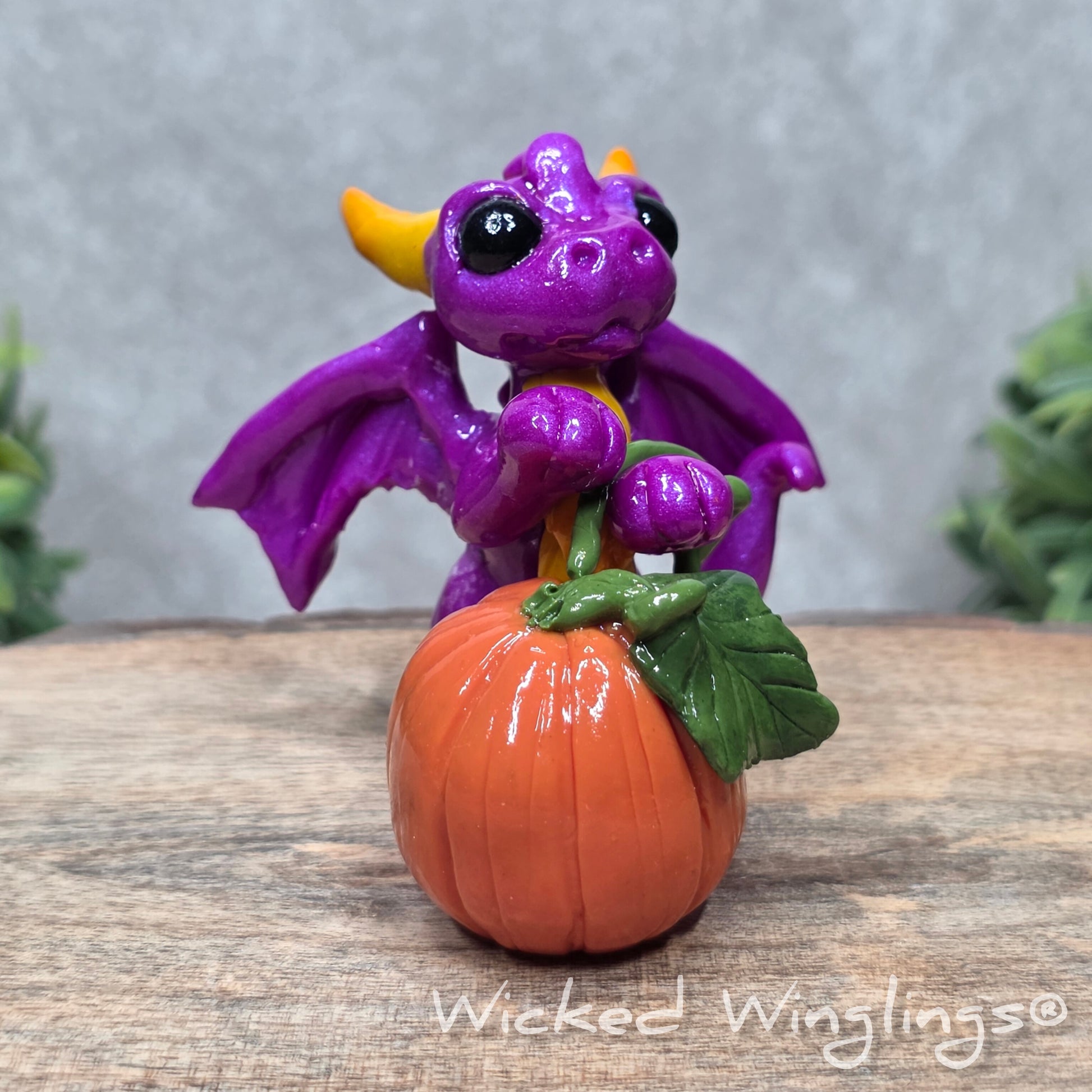 A baby clay dragon sculpture with a pumpkin against a wood background with greenery and text Wicked Winglings
