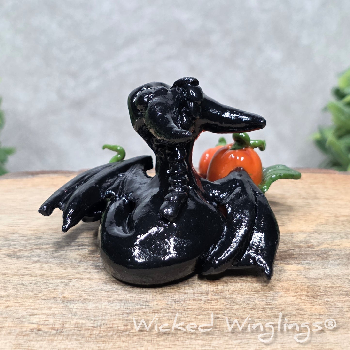 A baby clay dragon sculpture with pumpkins against a wood background with greenery and text Wicked Winglings