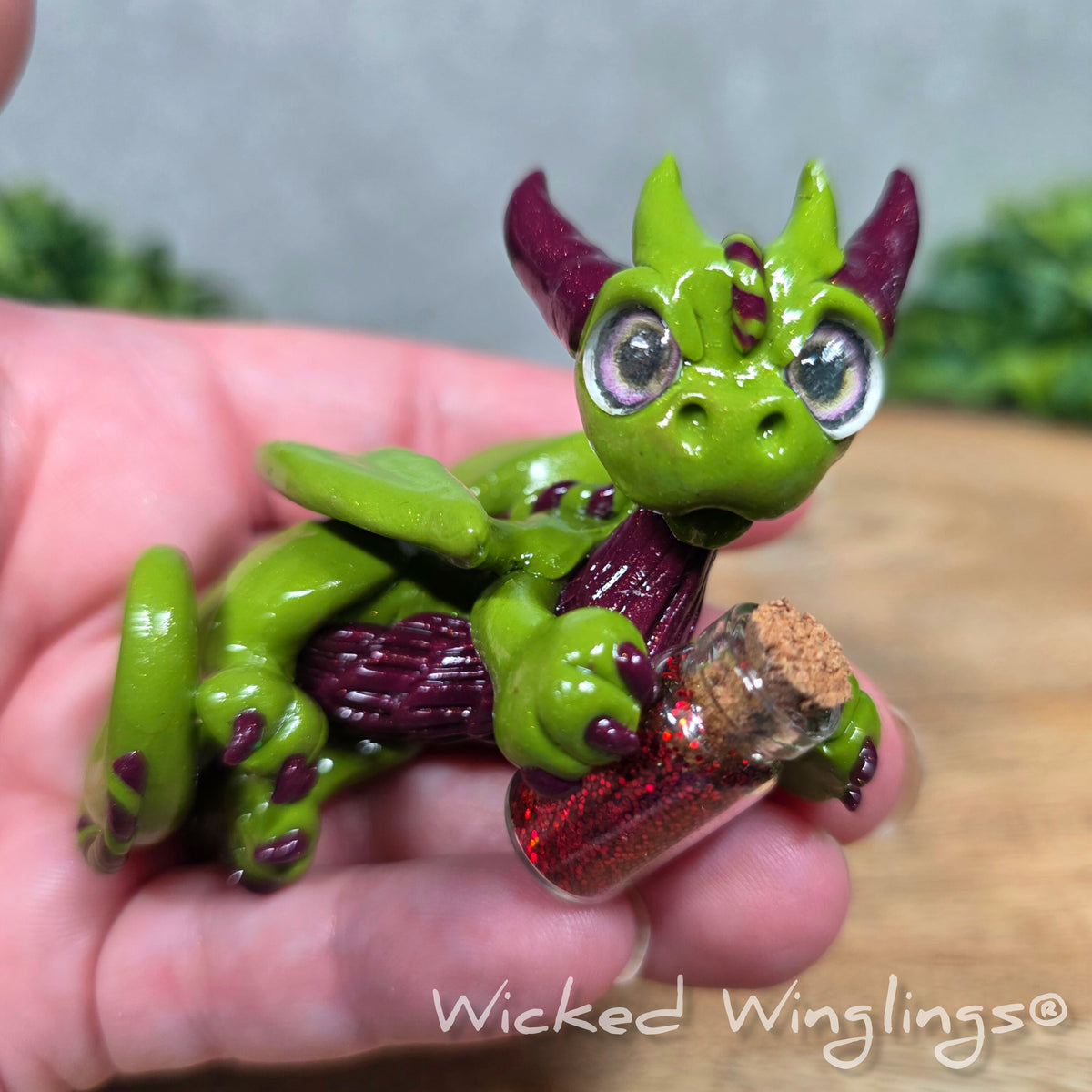 A hand holding a clay dragon with a potion bottle on a wood surface. Text reads Wicked Winglings.