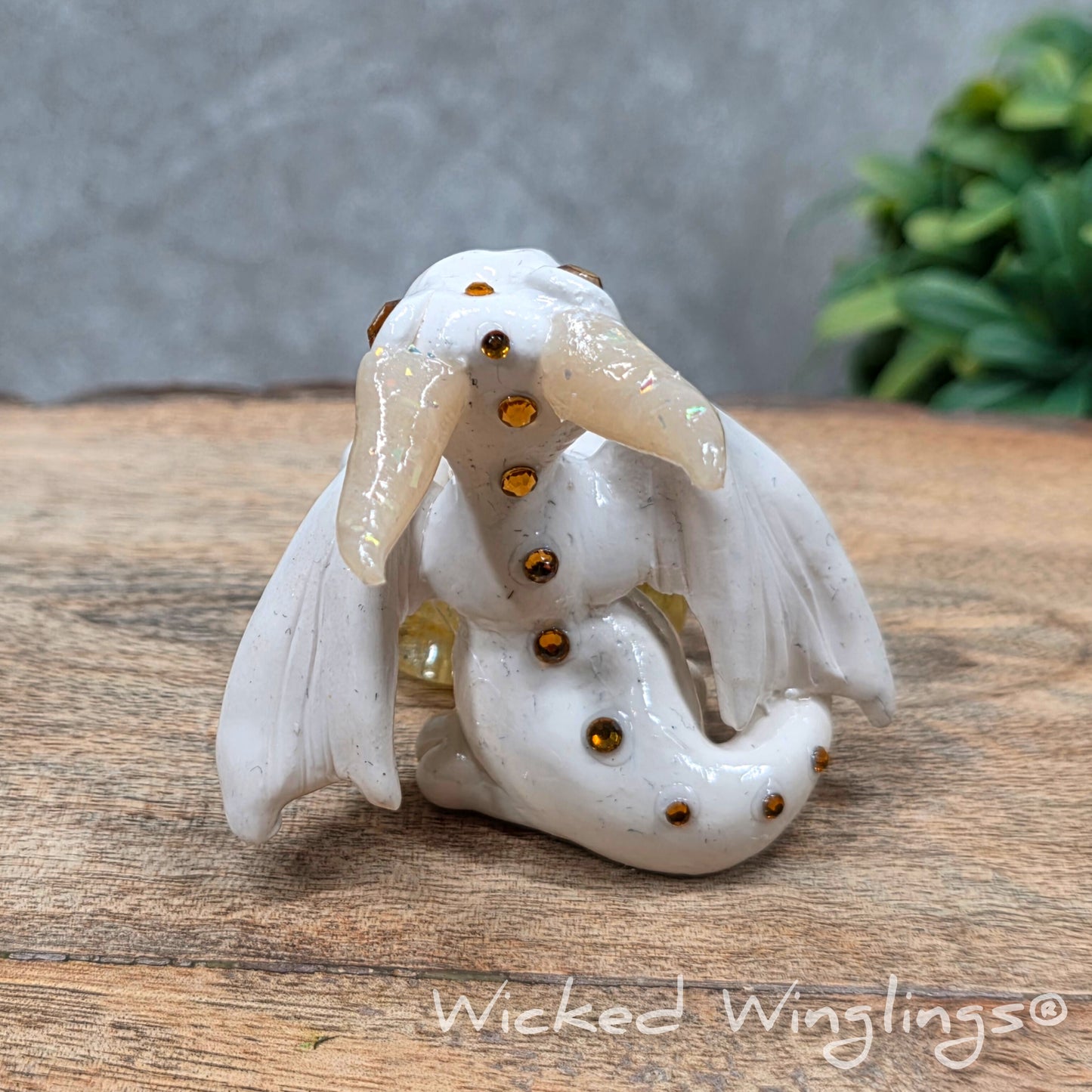 A baby clay dragon sculpture with a crackled quartz against a wood background with greenery and text Wicked Winglings