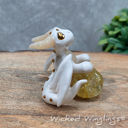 A baby clay dragon sculpture with a crackled quartz against a wood background with greenery and text Wicked Winglings