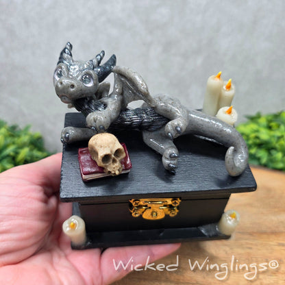 A hand holding a clay dragon on a black box with candles and a skull on a wood surface with greenery. Includes text Wicked Winglings
