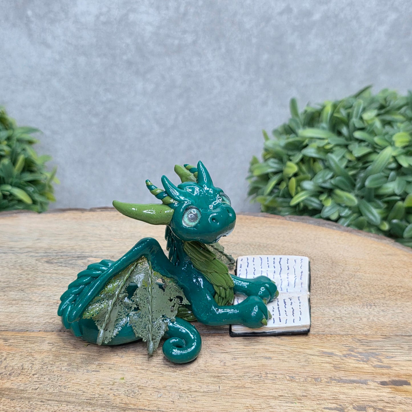 A dragon reading a book against a wood surface with greenery