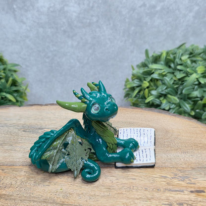 A dragon reading a book against a wood surface with greenery