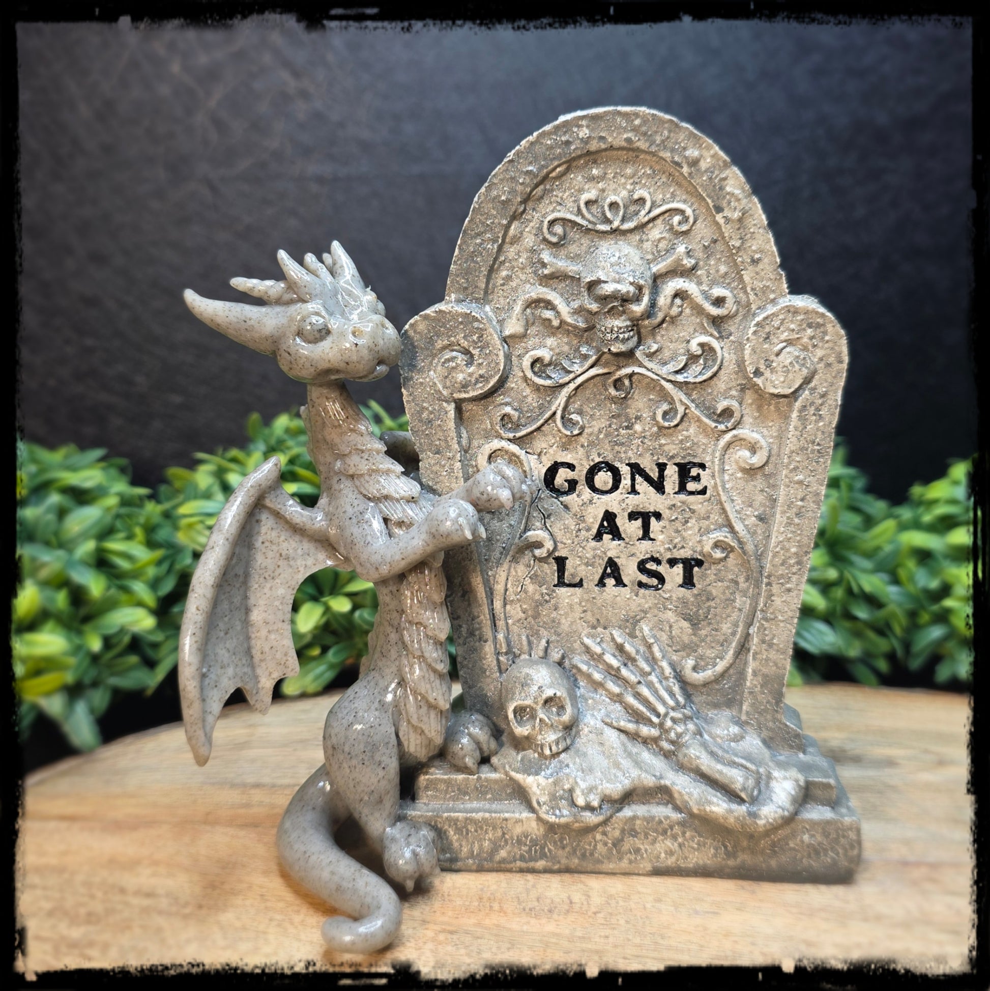 Decorative stone sculpture of a dragon and tombstone with 'Gone at Last' text, set against a blurred natural background.