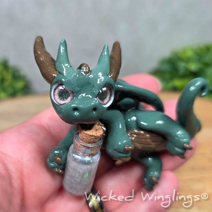 A hand holding a clay dragon with a potion bottle on a wood surface. Text reads Wicked Winglings.