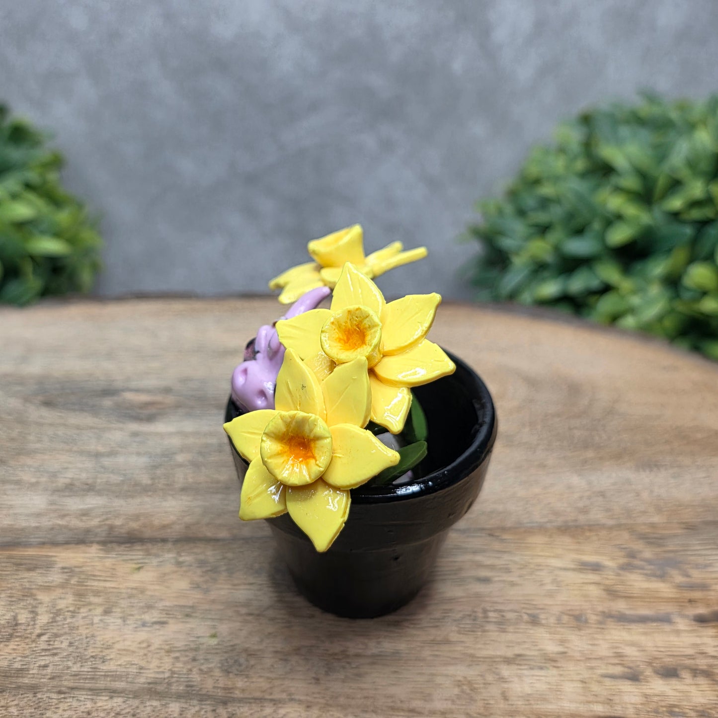 Side view of a clay baby dragon in a pot with daffodils against a wood surface with greenery.