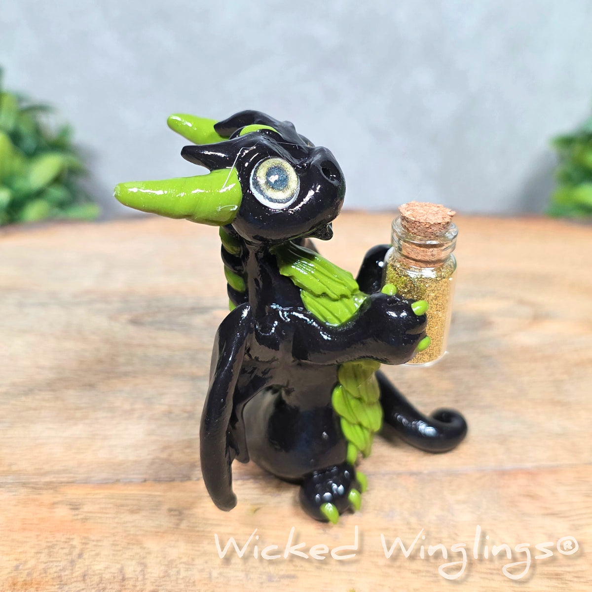 A clay dragon with a potion bottle on a wood surface. Text reads Wicked Winglings.