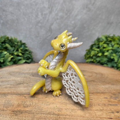 A clay dragon sculpture with lacy wings on a wood surface with greenery.