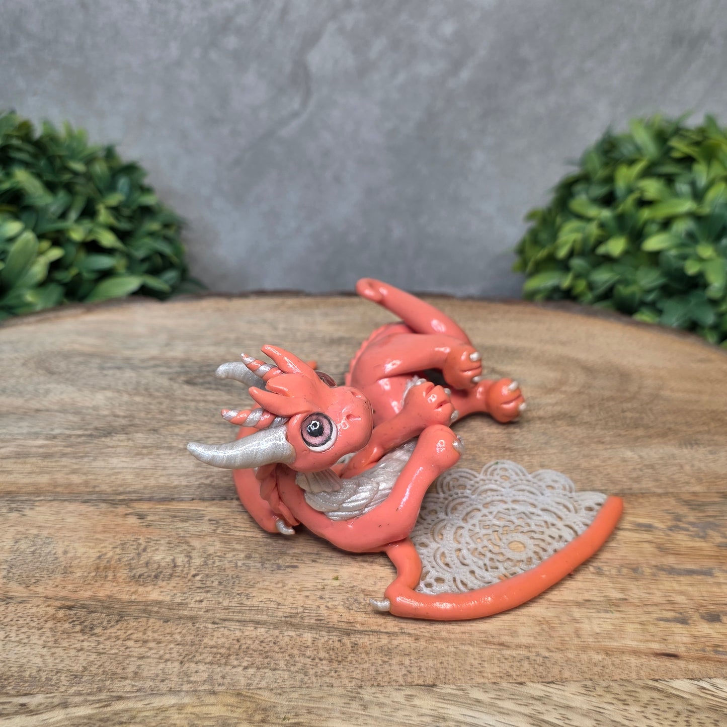 A clay dragon sculpture with lacy wings on a wood surface with greenery.