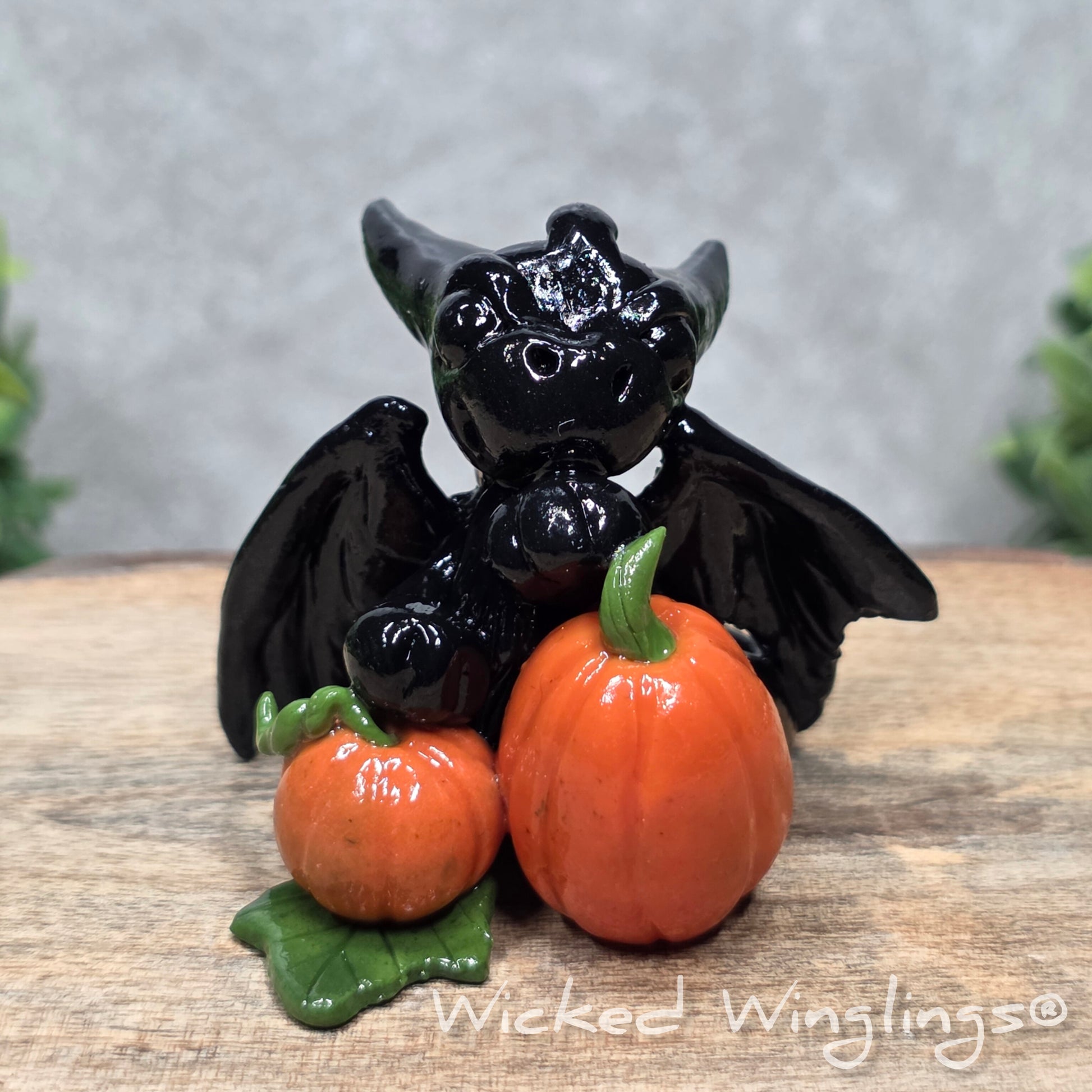 A baby clay dragon sculpture with pumpkins against a wood background with greenery and text Wicked Winglings