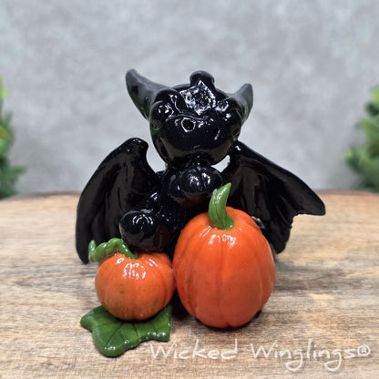 A baby clay dragon sculpture with pumpkins against a wood background with greenery and text Wicked Winglings