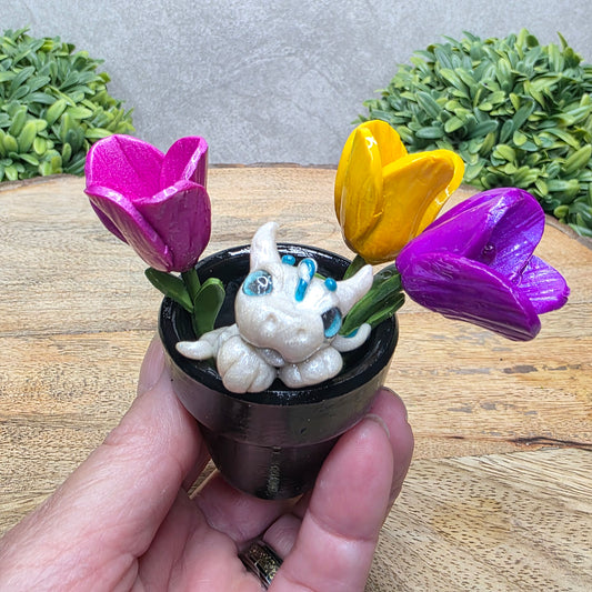 Hand holding a clay baby dragon in a pot with tulips against a wood surface with greenery.