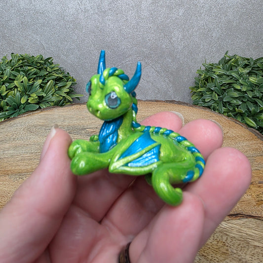 A hand holding a baby clay dragon against a wood background with greenery