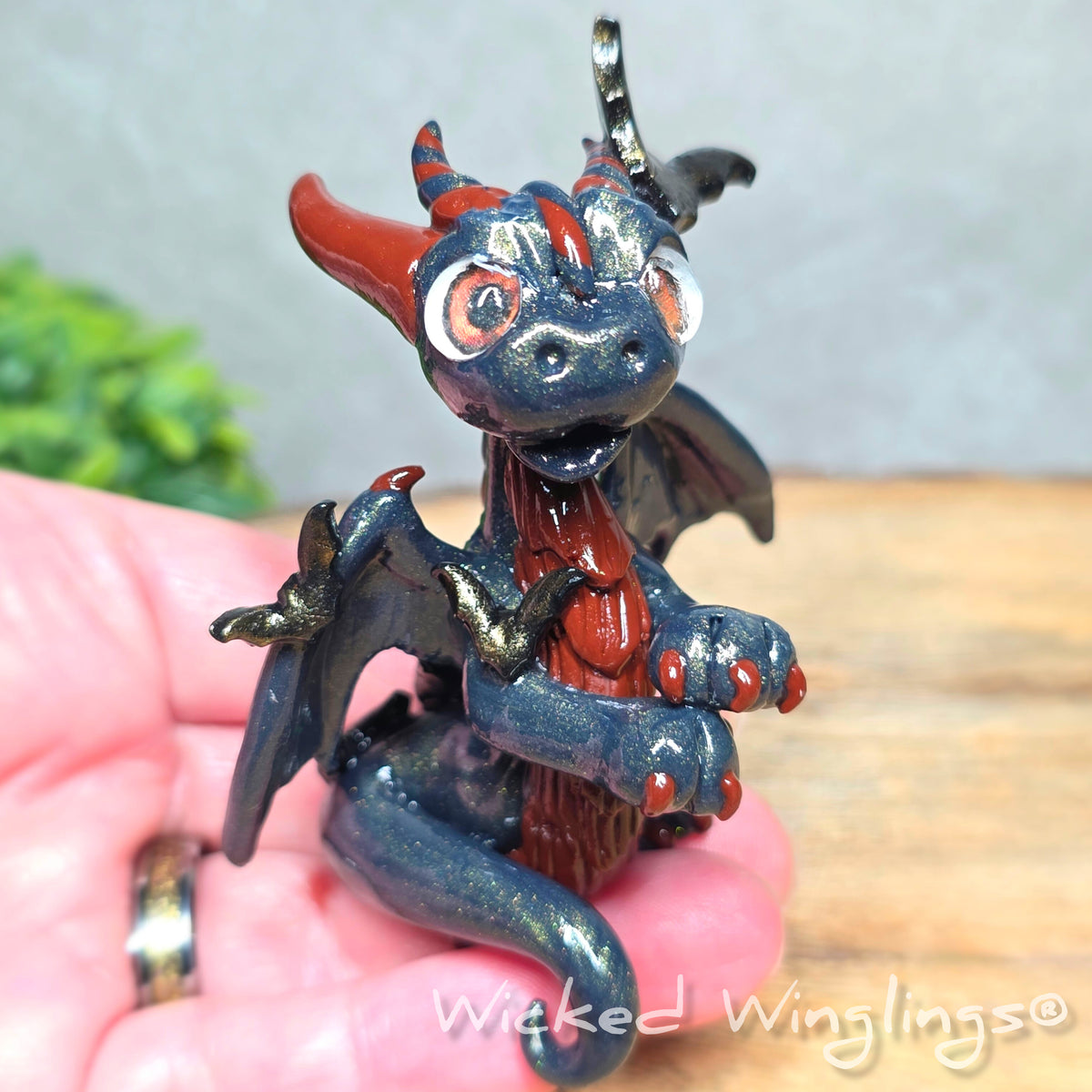 A hand holding a clay dragon with bats against a wood surface with greenery. Text reads Wicked Winglings.