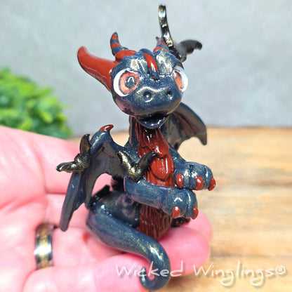 A hand holding a clay dragon with bats against a wood surface with greenery. Text reads Wicked Winglings.