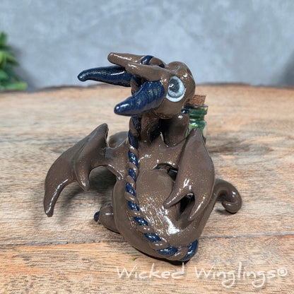 A clay dragon with a potion bottle on a wood surface. Text reads Wicked Winglings.