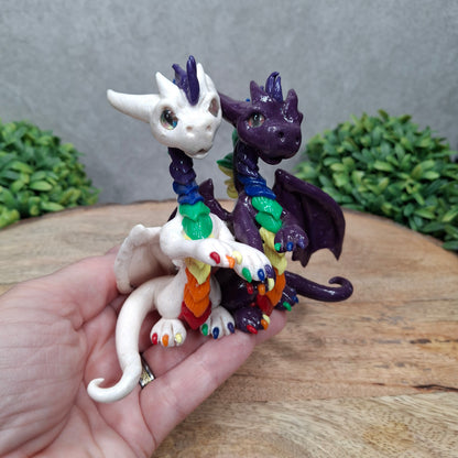A hand holding a dragon pair with rainbow accents against a wood surface with greenery