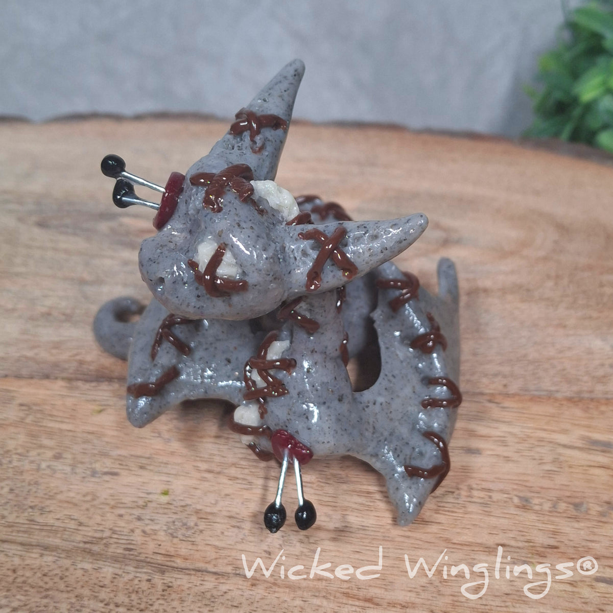 A clay voodoo dragon on a wood surface with greenery. Text says Wicked Winglings