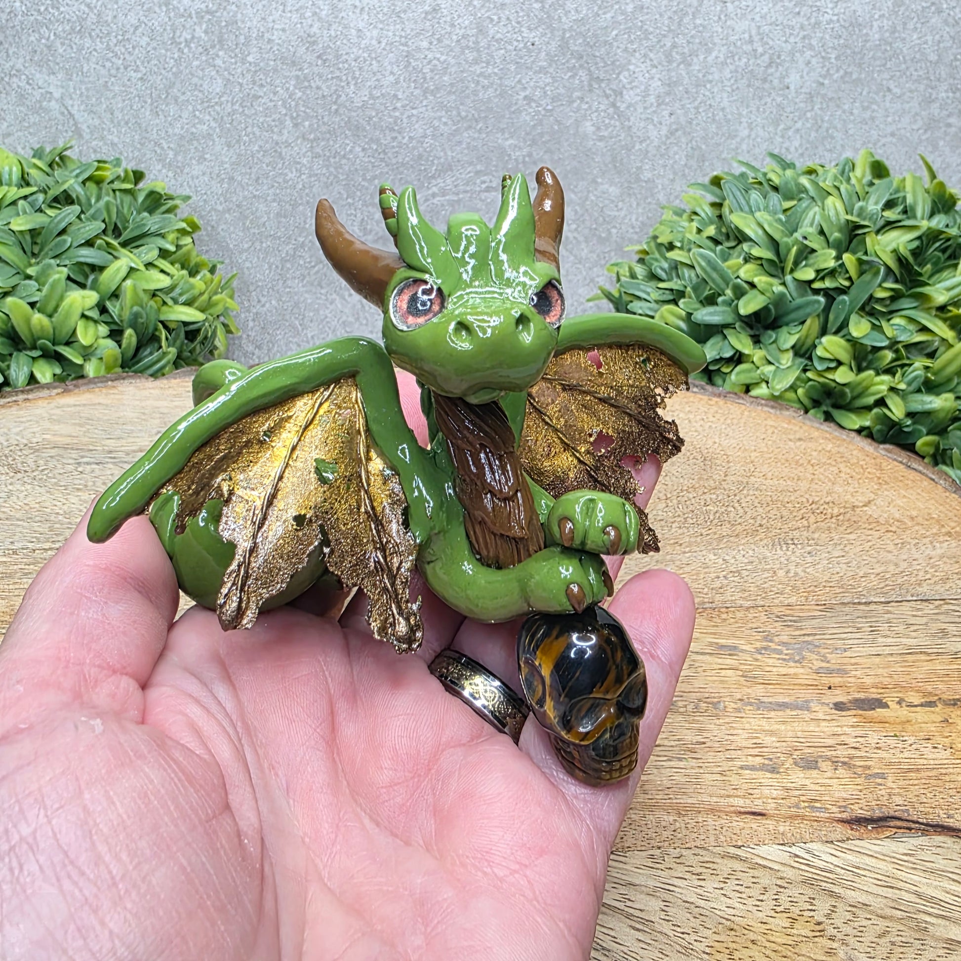Hand holding a clay dragon with a tigers eye crystal skull against a wood surface with greenery.